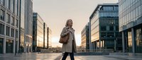 Woman in casual business attire standing in modern urban plaza at sunset, surrounded by contemporary glass and steel skyscrapers, ideal for corporate lifestyle and city living concepts.
