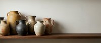 Ancient ceramic vessels and clay pots arranged on wooden shelf against neutral wall, suitable for home decor, interior design, and historical themed projects.