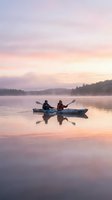 Two people kayaking on calm lake at sunrise with misty forested shoreline, peaceful morning water sports and outdoor recreation scene.