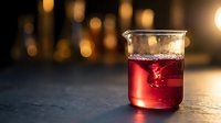 Red liquid in glass beaker on dark surface with warm bokeh lights creating atmospheric laboratory or bar setting for scientific research concepts.