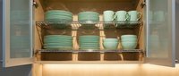 Organized kitchen cabinet with mint green ceramic dishes, bowls, and cups neatly stacked on wire shelves with warm LED lighting.