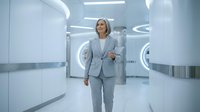 Middle-aged woman with blonde hair wearing a light gray business suit walking through a modern, minimalist corridor with white walls and contemporary lighting. She appears confident and professional, suitable for corporate, business, and leadership concepts.