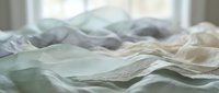 Unmade bed with rumpled white and green bedding sheets creating soft waves and textures in natural morning light for bedroom interior design concepts.