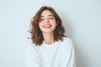 Young Asian woman with brown wavy hair laughing with closed eyes, wearing white sweater against light background. Natural joyful expression showing genuine happiness.