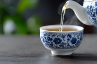 Steaming hot green tea being poured from traditional blue and white porcelain teapot into matching ceramic cup on wooden table with blurred background.