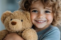 Happy young child with curly hair hugging brown teddy bear, smiling warmly at camera in cozy indoor setting for childhood and comfort concepts.