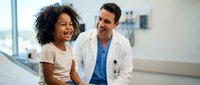 Happy African American girl laughing with friendly Caucasian male doctor in modern hospital corridor during pediatric consultation visit.