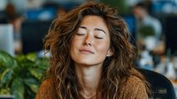 Young Caucasian woman with curly brown hair meditating peacefully with closed eyes in modern office environment, expressing mindfulness and workplace wellness.