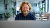 Middle-aged Caucasian businessman with curly hair and beard smiling at laptop in modern office workspace for corporate communications and remote work.