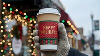 Gloved hand holding festive coffee cup with happy holidays message against blurred winter street decorated with colorful lights and seasonal decorations.