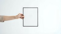 Hand holding blank white picture frame against neutral wall background for mockup and presentation purposes.