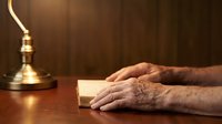 Elderly hands resting on open book beside vintage brass lamp on wooden desk, symbolizing wisdom, knowledge, and lifelong learning in warm study setting.