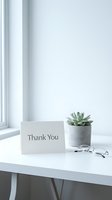 Minimalist white desk with a thank you card, succulent plant in concrete pot, and glasses by a bright window.