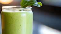 Fresh green smoothie with spinach leaf garnish in clear glass against dark background, healthy breakfast drink rich in vitamins and nutrients.