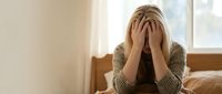 Stressed Caucasian woman sitting on bed covering face with hands, expressing anxiety, depression, or emotional exhaustion in a bedroom.