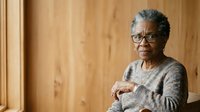Senior African American woman with gray hair and glasses wearing a gray cardigan, standing confidently against a wooden wall background for portrait and lifestyle use.