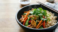 Asian stir-fry with mixed vegetables, mushrooms and brown rice in black bowl with chopsticks on wooden table for healthy meal preparation.
