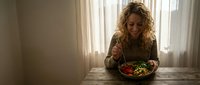 Young blonde Caucasian woman enjoying healthy salad bowl at wooden table by window in cozy home kitchen during daytime meal.