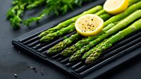 Fresh green asparagus spears on black grilling pan with lemon halves and peppercorns, dark moody background with herbs, ready for cooking healthy meal.