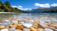 Smooth river rocks and pebbles in clear shallow water with mountain lake and snow-capped peaks in background, low angle view capturing tranquil wilderness landscape.