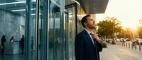 Confident businessman in suit standing outside modern glass office building during golden hour sunset, professional corporate lifestyle and success concept.