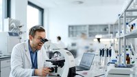 Male scientist of South Asian descent wearing white lab coat and glasses, examining samples under microscope in modern laboratory setting with equipment and colleagues in background, suitable for research, healthcare, education and biotechnology content.