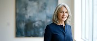 Professional mature Caucasian businesswoman with gray hair in navy blazer standing confidently by office window with abstract art background.