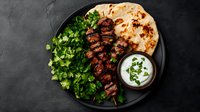 Grilled meat kebab skewers served with fresh cilantro, flatbread and yogurt sauce on black plate. Middle Eastern cuisine photographed from above.