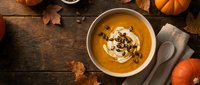 Creamy pumpkin soup with whipped cream and spices in white bowl on rustic wooden table with autumn leaves and orange pumpkins for seasonal cooking.