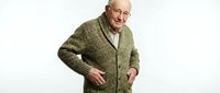 Senior man with glasses wearing olive green cardigan sweater, smiling at camera against white background. Suitable for healthcare, lifestyle, and senior living content.