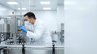 Male scientist in white lab coat and protective mask operating pharmaceutical manufacturing equipment in sterile production facility.