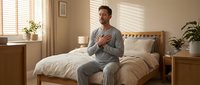 Middle-aged Caucasian man in casual gray pajamas sitting on wooden bed in bright, minimalist bedroom with potted plants and natural light from windows, suitable for wellness and sleep content.