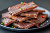 Spicy beef jerky strips with chili flakes and herbs on dark plate. High-protein snack food for healthy eating and outdoor adventures.
