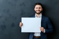 Young Caucasian businessman in navy suit holding blank white paper against dark wall background, displaying confident smile and professional demeanor.