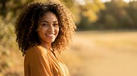 Young African American woman with curly hair smiling outdoors in golden sunlight wearing casual clothing for lifestyle and wellness concepts.