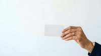Adult male hand with medium skin tone holding blank white business card against neutral background for corporate branding and professional marketing purposes.