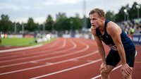 Male athlete in dark athletic wear bent forward in ready position on red running track with blurred spectators and green trees in background during outdoor track and field competition event.