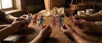 Diverse paper cutout figures held by multiple hands in warm indoor lighting, representing family unity, teamwork, community support, and togetherness for social concepts.