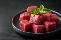 Fresh raw beef cubes garnished with basil leaves on black ceramic plate against dark background. Premium quality meat for cooking and restaurant menu.