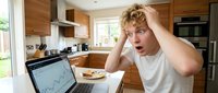 Shocked young man with curly blonde hair and fair complexion sits at kitchen table with laptop showing financial charts, expressing surprise or concern about stock market performance or investment results.