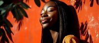 Young African American woman with long dark hair smiling with eyes closed against vibrant orange wall, wearing yellow top, with tropical leaves creating natural shadow patterns.