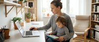 Young mother working from home on laptop while caring for toddler daughter, multitasking parent balancing career and childcare in modern living room.