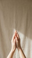 Caucasian hands pressed together in prayer gesture raised upward against soft beige linen background with gentle natural light.