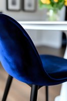 Modern dining chair upholstered in rich royal blue velvet fabric with curved backrest and black metal legs, shown in close-up against blurred interior background.
