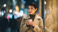 Young Asian woman in beige coat holding coffee cup on city street at night with bokeh lights background for lifestyle and urban concepts.