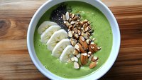 Green smoothie bowl topped with sliced banana, mixed nuts, chia seeds on wooden table. Healthy breakfast concept for clean eating and detox.