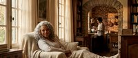 Elderly woman with curly gray hair relaxing on beige sofa in cozy library room with brick fireplace and wooden bookshelves during daytime.