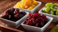 Fresh and dried fruits served in white ceramic bowls on wooden table, including dates, cranberries, oranges, and kiwi slices arranged in modern presentation.
