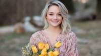 Young Caucasian woman with blonde wavy hair wearing floral pink dress holds bouquet of fresh yellow tulips and white wildflowers, smiling outdoors in spring.