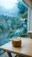 Cozy cafe scene with a glass mug of latte on a wooden table, Welcome written on a foggy window, green plants visible outside on a rainy day.
